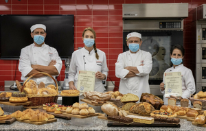 Four bakers in white Richemont master baker uniforms and certificates standing behind a countertop filled with assorted baked goods in a kitchen