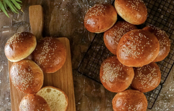 Freshly baked sesame hamburger buns on a wooden board and cooling rack.
